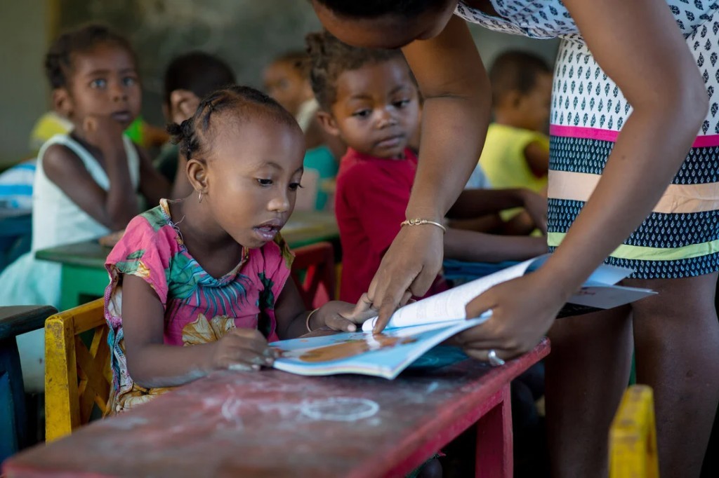 Teacher helping a young girl read at a worn classroom desk